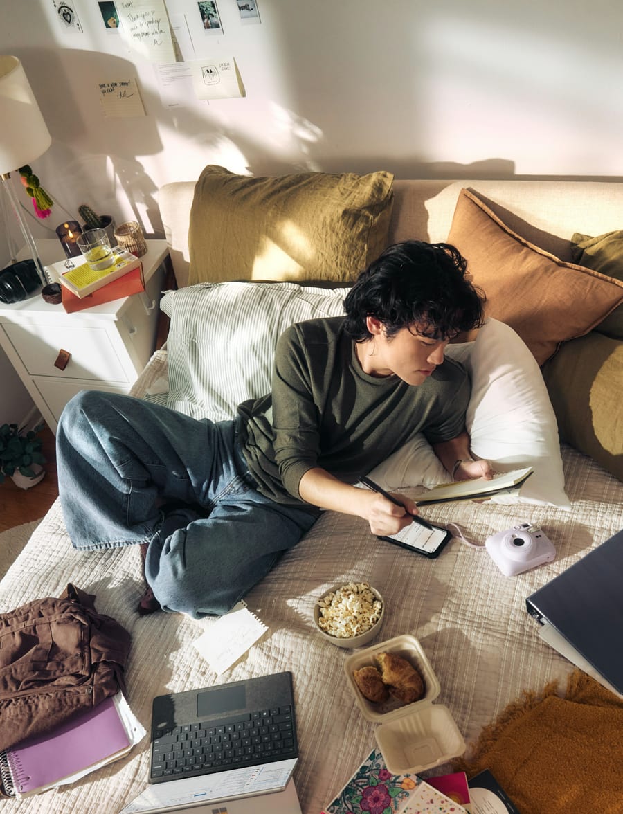 A person lying on a bed drawing on a tablet, surrounded by their things